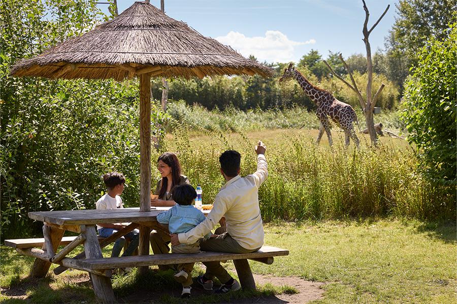 Gezin aan een picknicktafel met rieten parasol en giraffe op de achtergrond